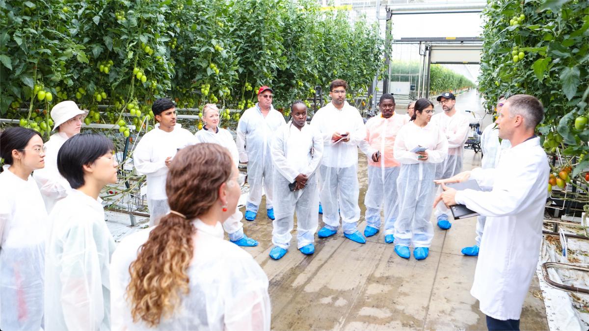 Group of students in white coveralls in a greenhouse listening to a professor