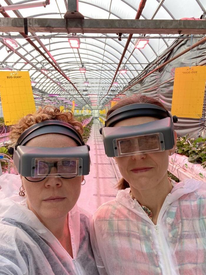 Cara McCreary and a colleague standing inside a greenhouse wearing coveralls and matching visors