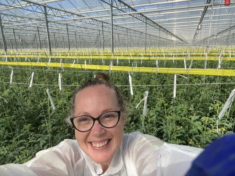 Cara McCreary smiling and standing infront of a greenhouse full of green plants 
