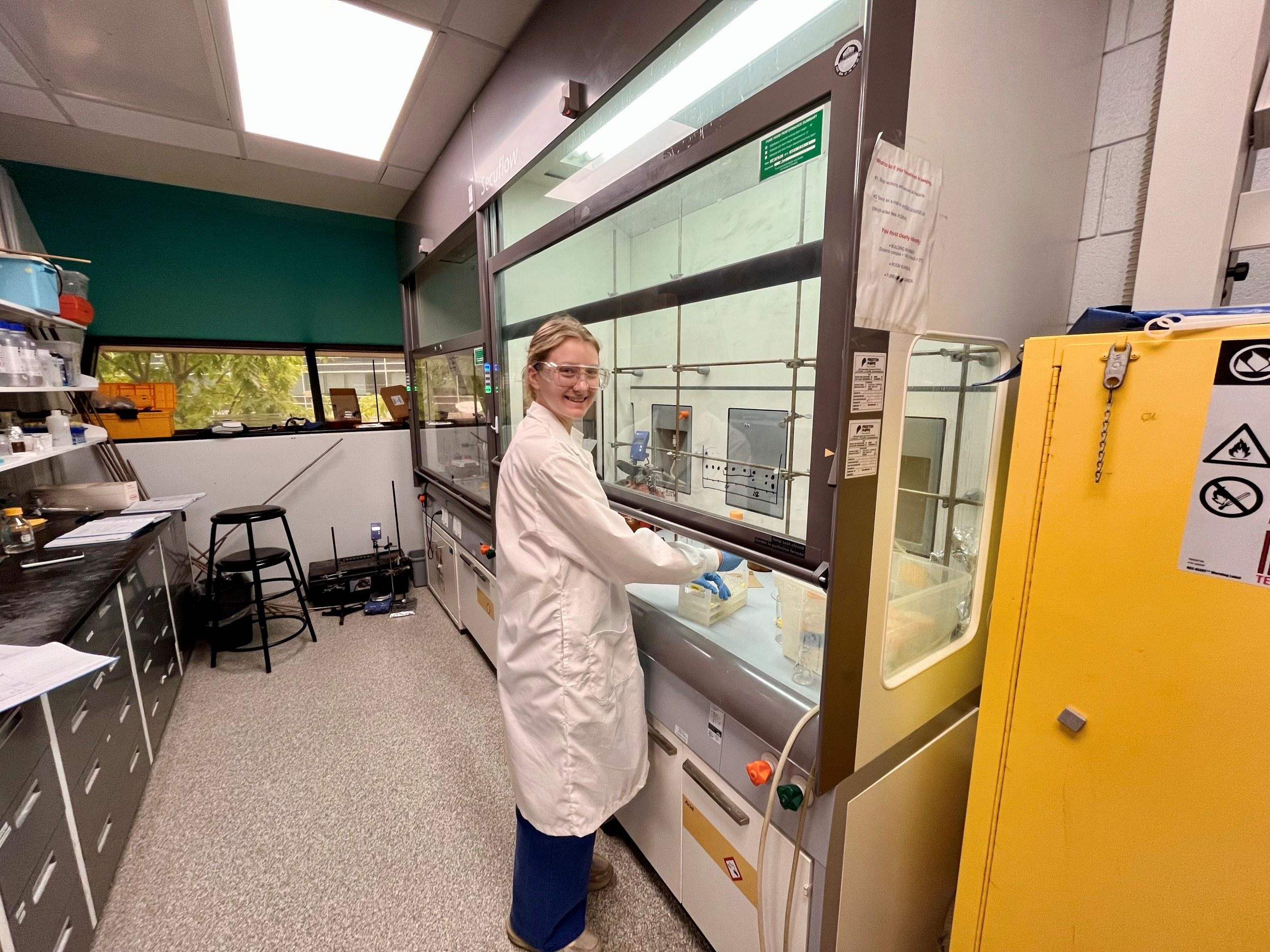 Lucy stands works in a fume hood while wearing a white lab coat and safety glasses.