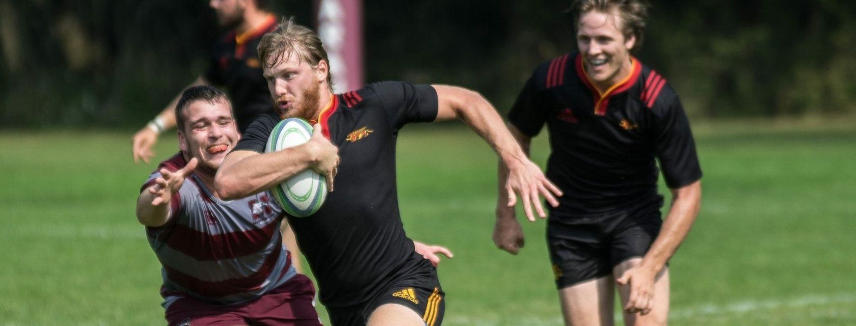 Guelph Grad Jordan Hofstra running across a rugby field while opposing players attempt to tackle him.