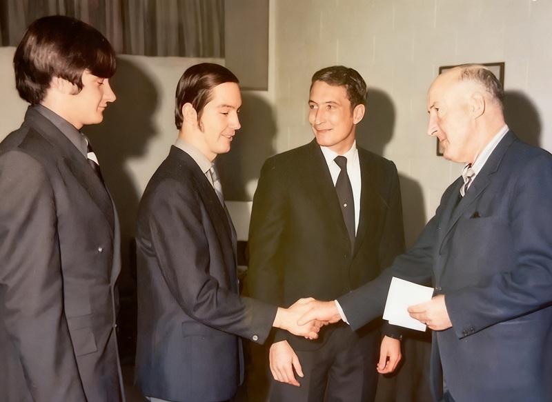 Three male students wearing suits standing in front of male instructor. One male student is shaking hands with instructor receiving an award.