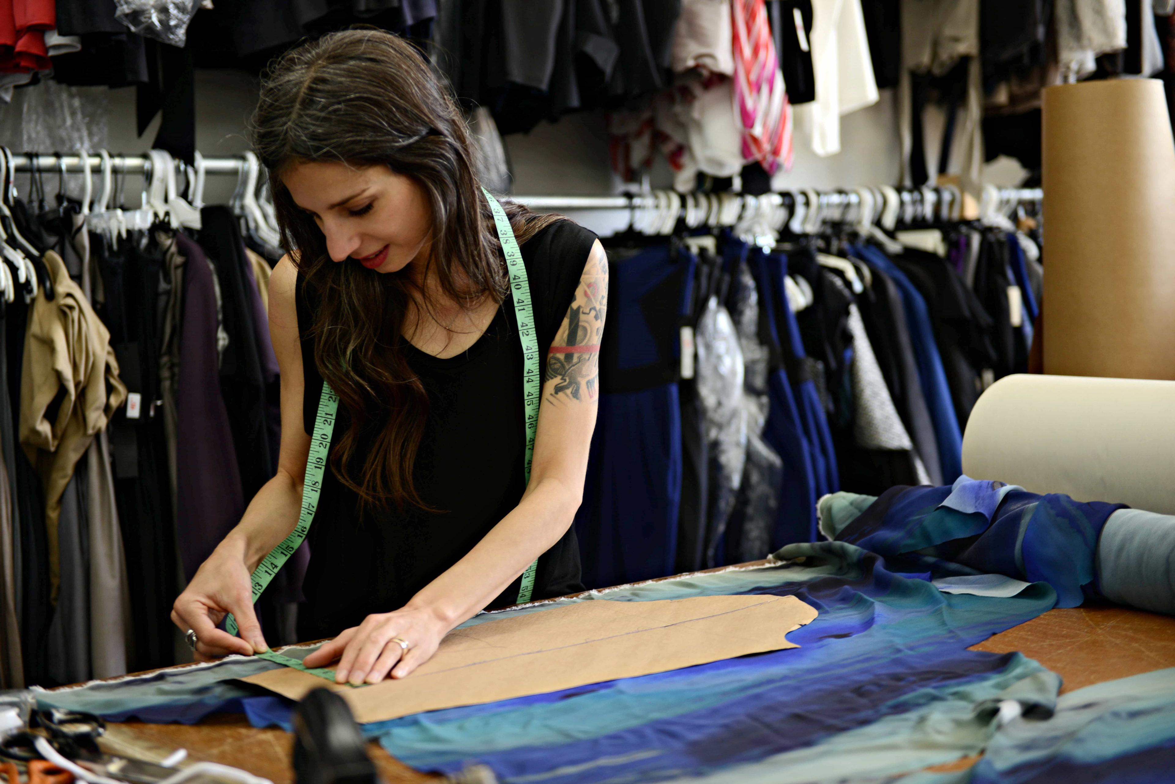 The late Valérie Dumaine is working at a table in her Montreal fashion design studio, surrounded by various garments on hangers. She is measuring fabric with a tape measure. Rolls of fabric are visible on the table she is working on.