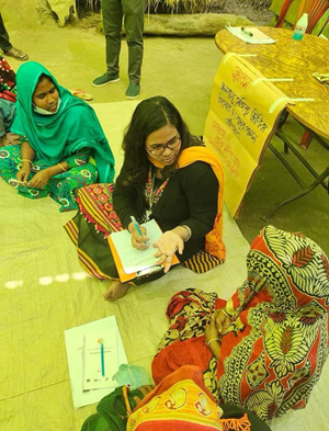 Person taking notes while speaking with community members seated on the floor during a food and community discussion session.