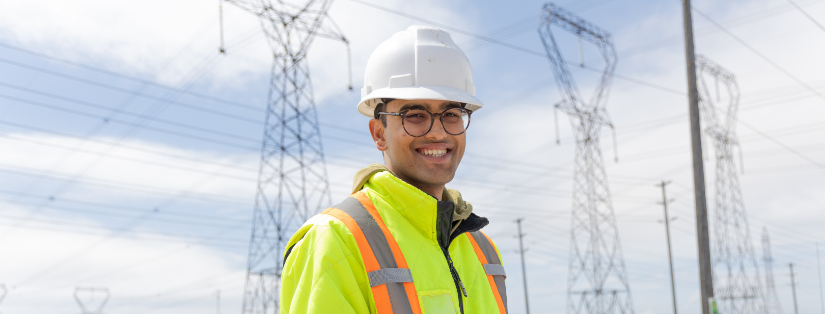 An engineering student wearing a hi-vis vest and white hard hat stands in front of an electrical corridor.