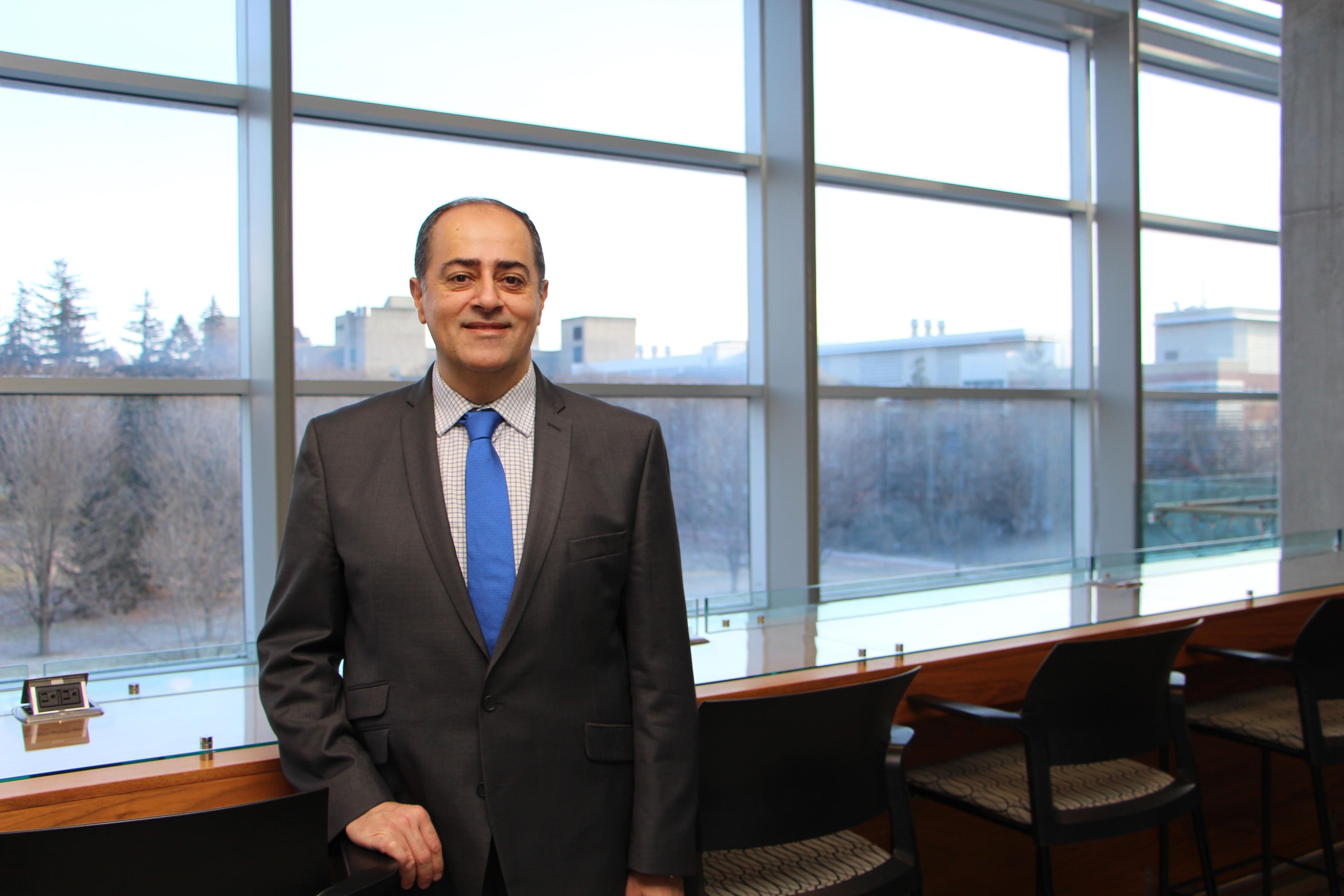 Dr. Shayan Sharif stands in front of a wall of windows overlooking the University of Guelph campus, smiling at the camera.