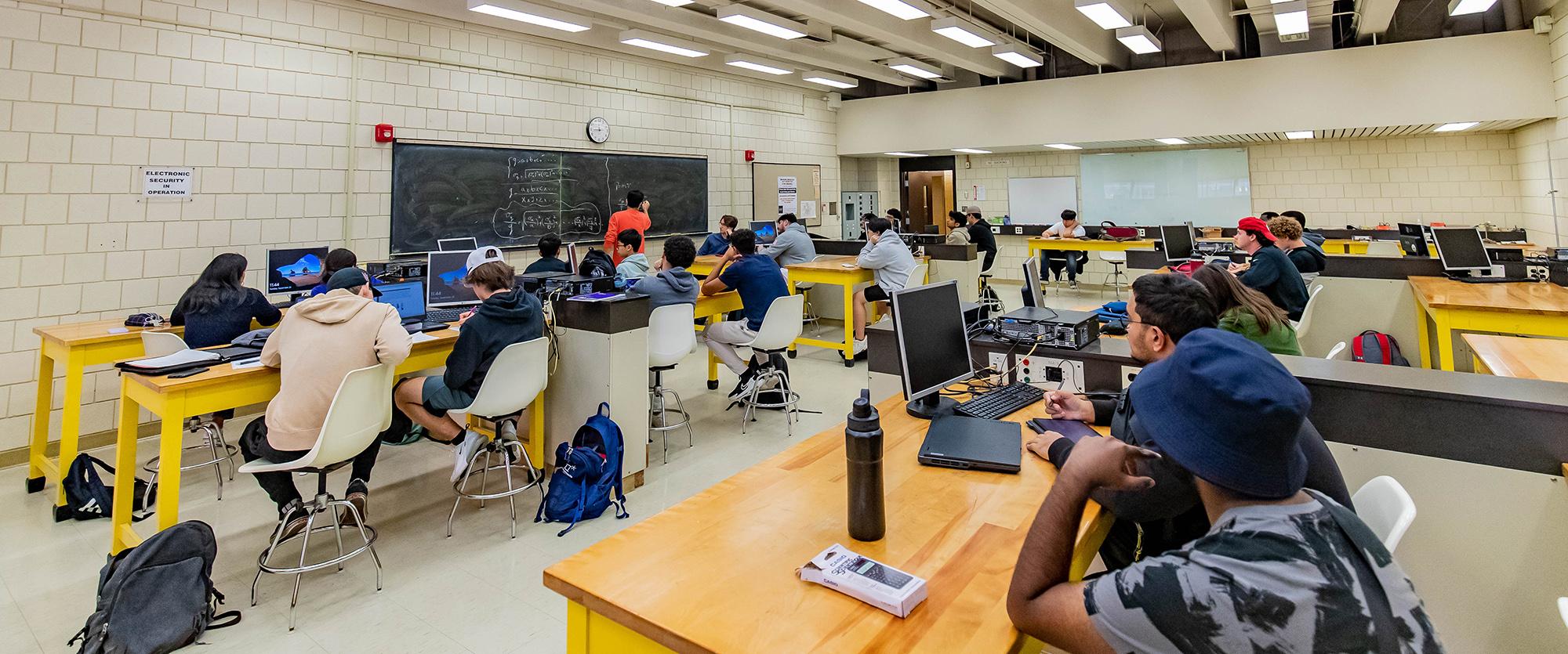 Students are in a physics lab being taught by a graduate teaching assistant, who is writing equations on a chalkboard while students observe.