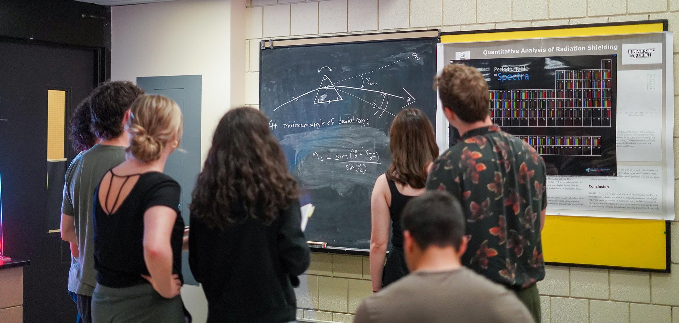 A group of students stand around a physics chalkboard at the university of guelph