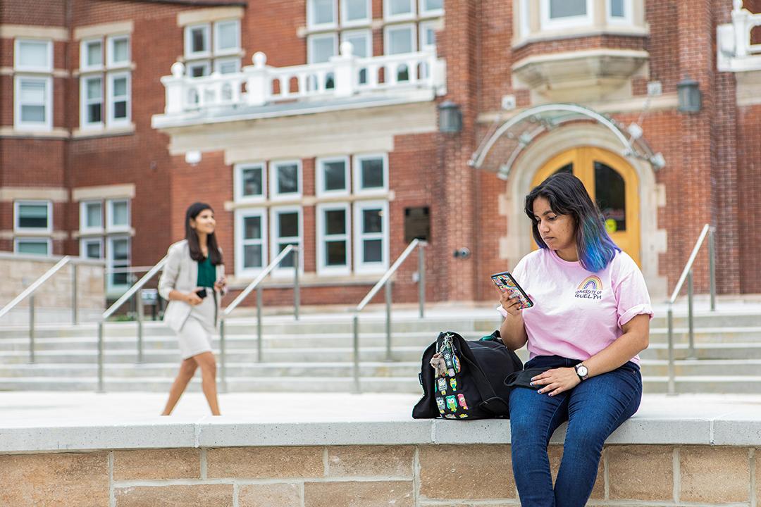 A student sits on their phone in the Lang Plaza at the University of Guelph while another student in a suit walks in the background.