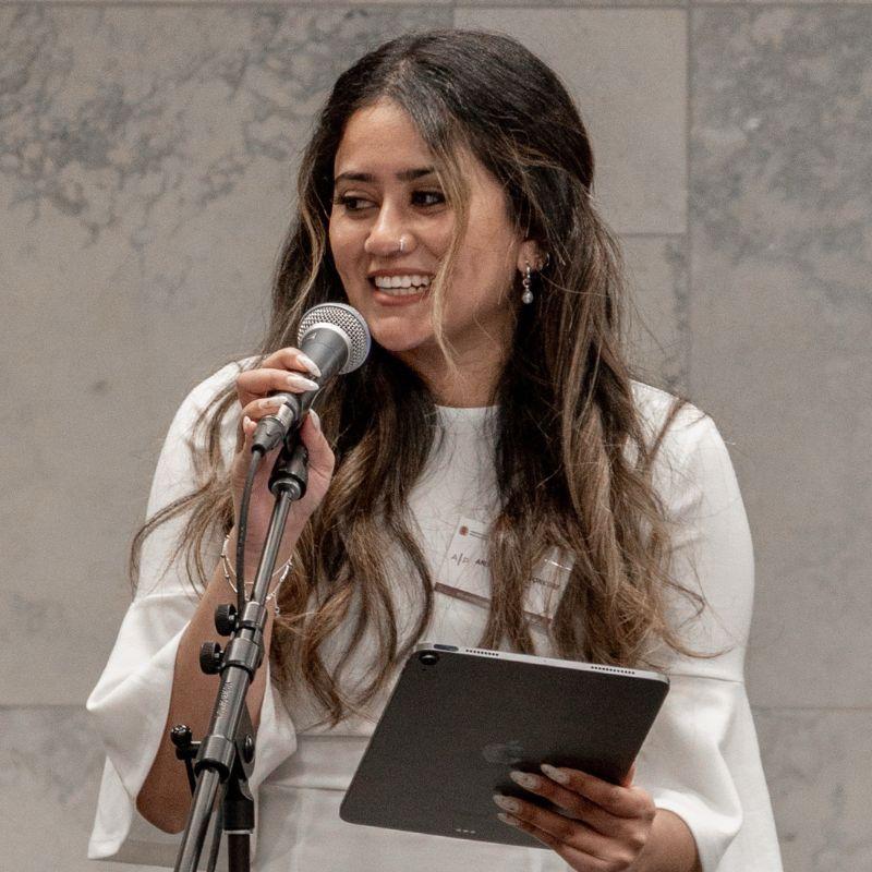 Anindita Ponkshe standing behind a microphone speaking at an event.