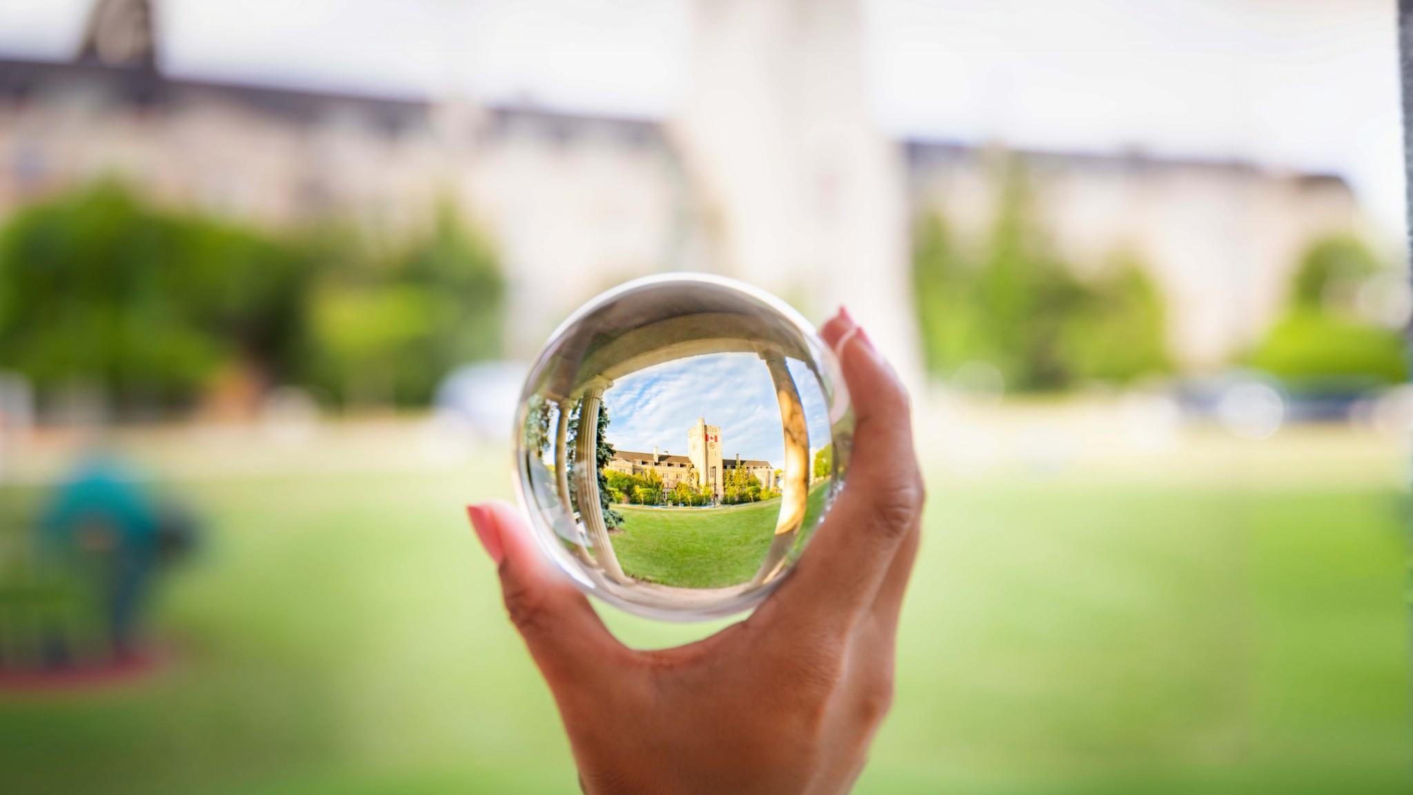A hand holding a clear glass sphere that shows a reflection of Johnston Hall
