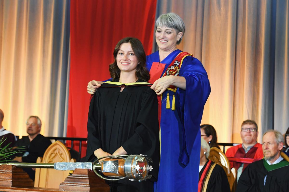 Julianna Tindall wearing a black gown and smiling as someone places a yellow hood on her gown