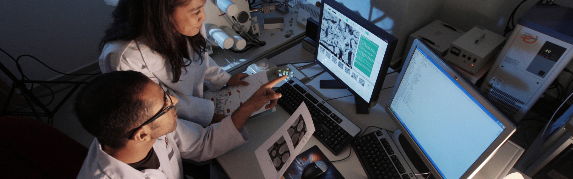 Two students in lab coats looking at data on two computer screens