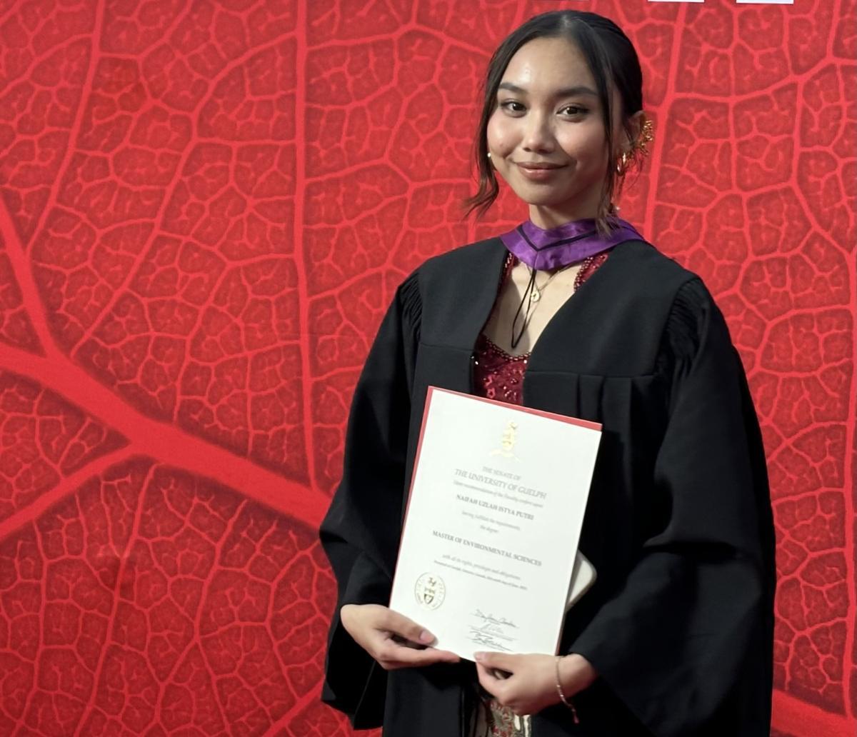 Student standing in front of red backdrop in convocation robes holding her degree