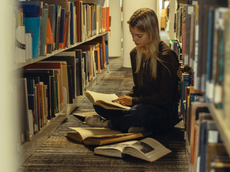 A student sits on the library floor, between the stacks. She reads a book, and several books are open around her on the floor.