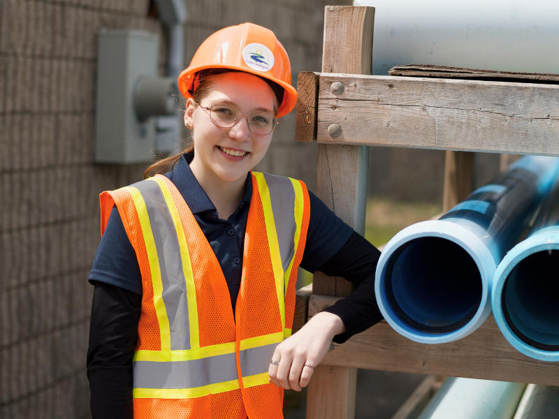 A student wearing a safety vest and hard hat poses next to city plumbing pipes.