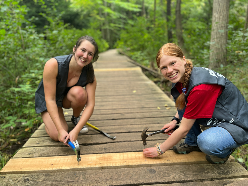 Two student volunteers use hammers to fix up a wooden boulevard running through a forest.