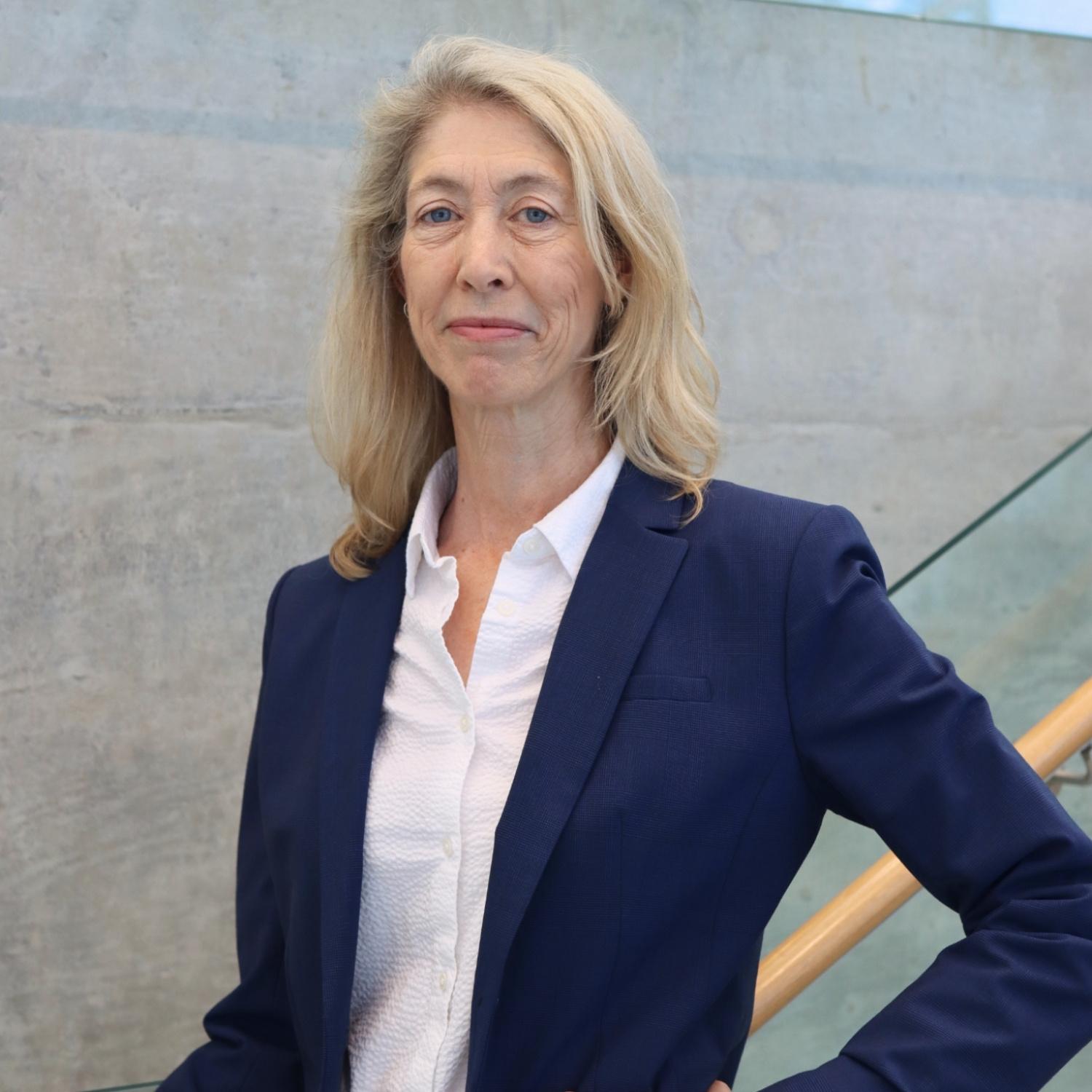 blonde caucasian woman wearing a navy blue blazer, posing with her hand on her hip for a headshot