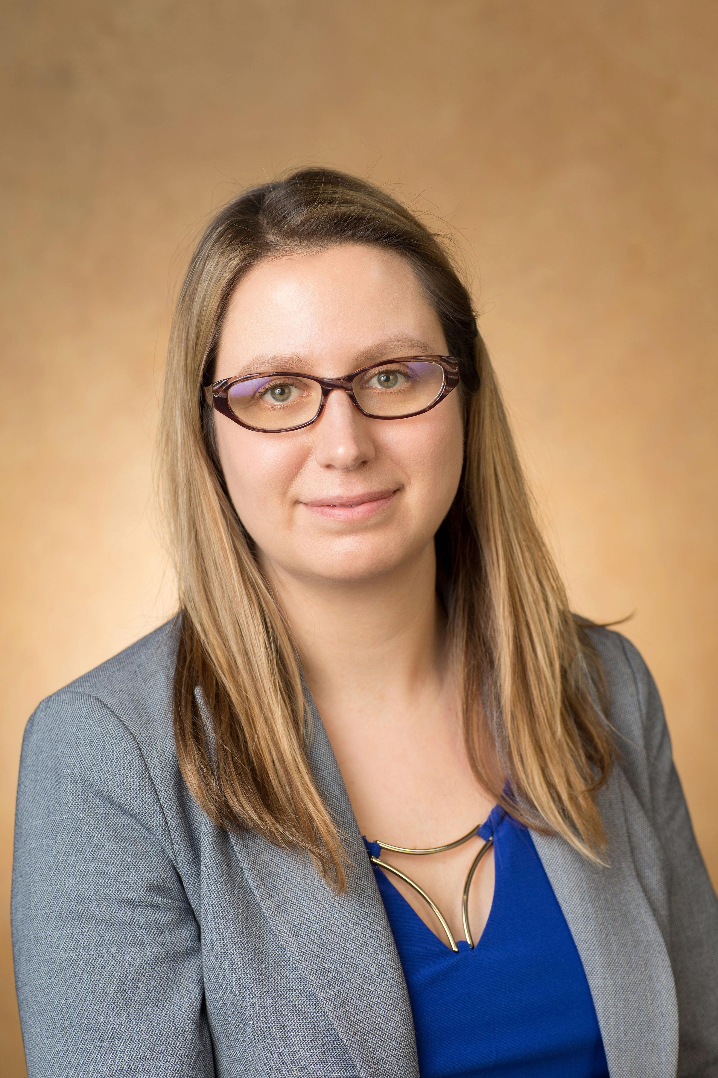 A professional headshot of Meaghan Hancock, a blonde fair-skinned woman with glasses wearing a grey blazer and blue shirt