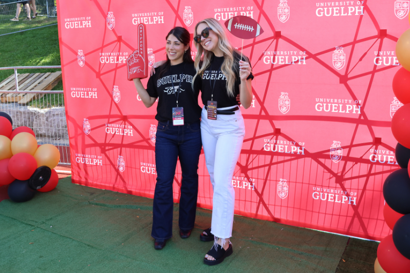 Two people pose for a picture in front of a red background that features University of Guelph branding. They are holding a foam finger and a cut-out of a football, while outdoors with balloons surrounding the facade.
