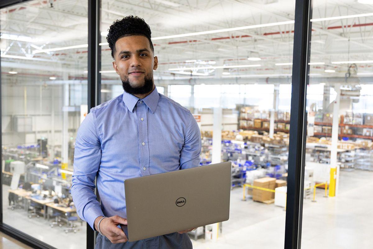 Experiential Learning Students stands holding a laptop with a window looking into a warehouse behind them