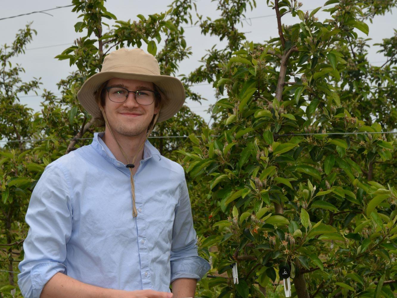 Owen wears glasses and a wide-brimmed hat, smiling in front of an apple tree