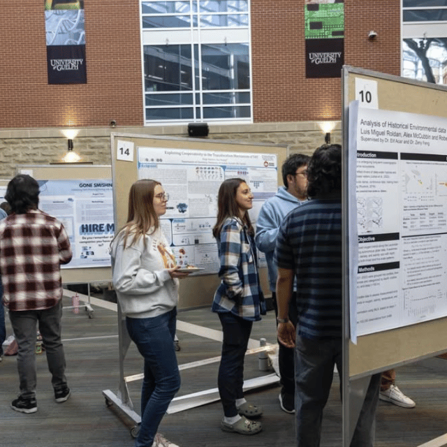 Students crowd around a bulletin board as a graduate student presents their research.