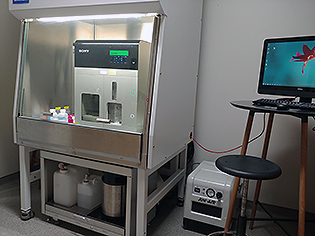 Picture of a Biosafety cabinet with the Sony Cell Sorter i it, also showing the compressor, the liquids cart of the instrument, and the computer