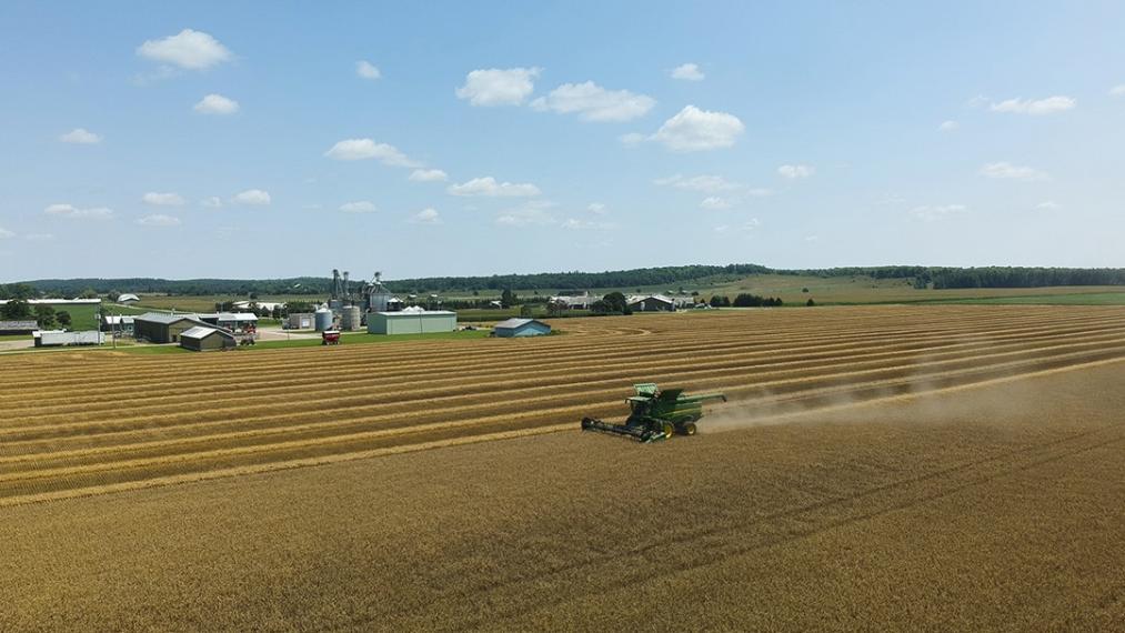 Aerial photo of wheat being harvested in a golden field with farm buildings in the background