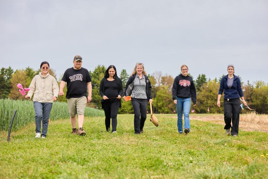 A line of six smiling researchers walks across a field of cut grass. One researcher holds a shovel in her hand.