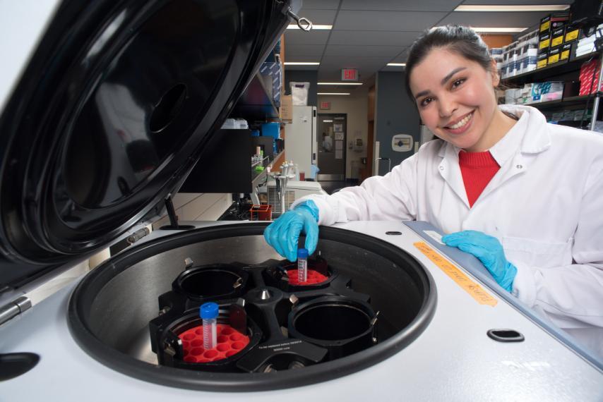 Dr. Fatemeh Fazel wears a lab coat and smiles, posing for a photo in front of a centrifuge machine in her lab
