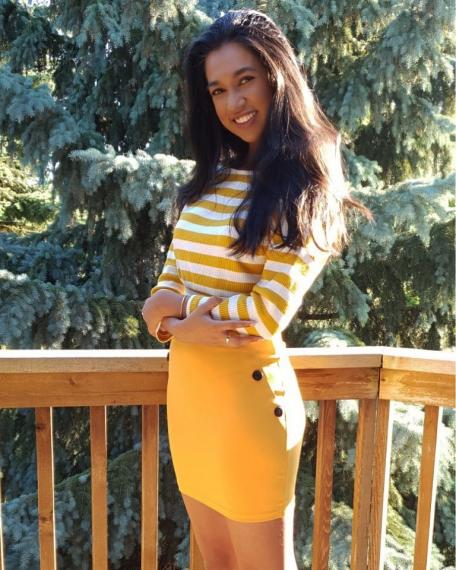 Photo of Maleeka Singh standing on a deck in front of a pine tree with her arms crossed.