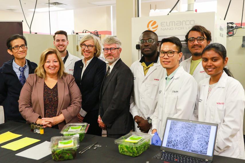 A group of people gather in a laboratory and smile for a photo op: OMAFRA Minister Lisa Thompson is flanked by Dr. Loong-Tak Lim and U of G President Charlotte Yates; Dr. Malcolm Campbell and five student researchers wearing lab coats are also present