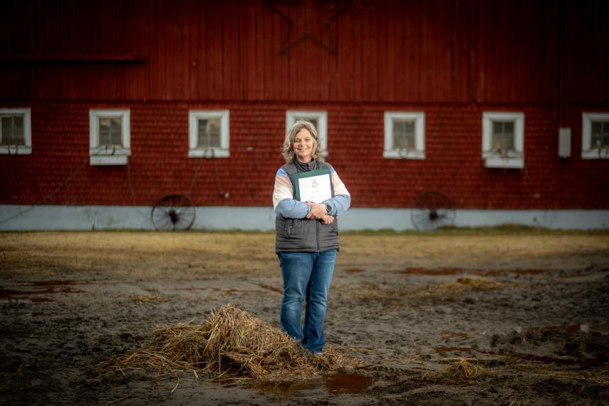 Kim Jo Bliss, a woman with grey hair and wearing blue jeans, stands in front of her barn holding a certificate to her chest