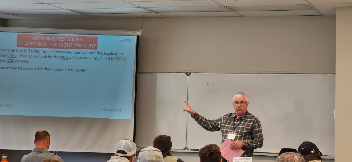 At the front of a classroom, an instructor gestures toward a projection