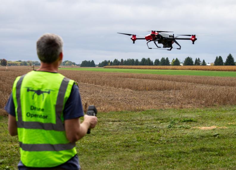 Ian is flying a drone over a farm field. He is wearing a bright yellow safety vest that says "Drone Operator"