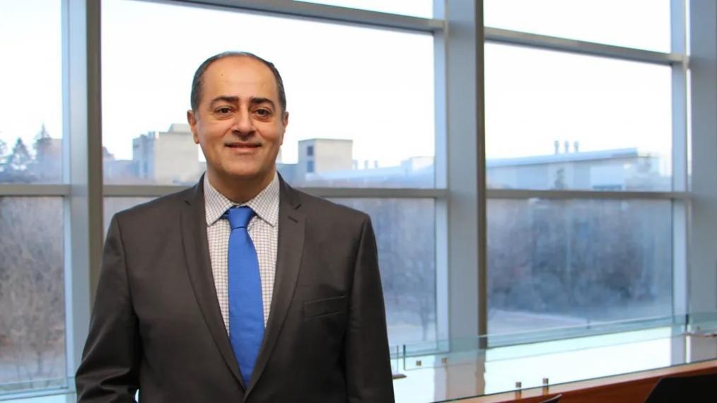 Dr. Shayan Sharif, wearing a suit and tie, stands in front of a scenic U of G window