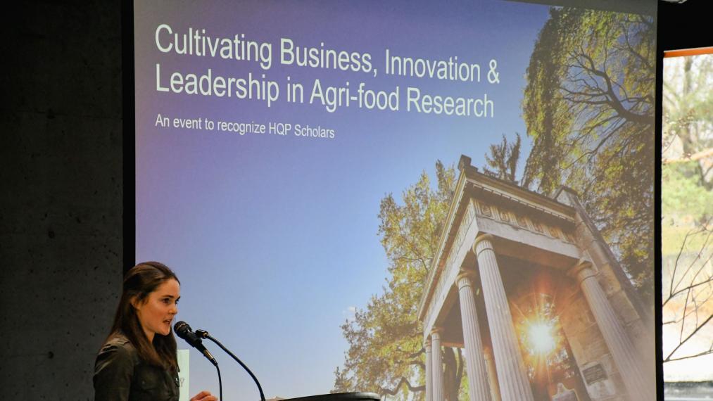 Hannah Woodhouse, a woman with long dark hair, speaks into a microphone in front of a PowerPoint presentation with the event title "Cultivating business, innovation and leadership in agri-food research"