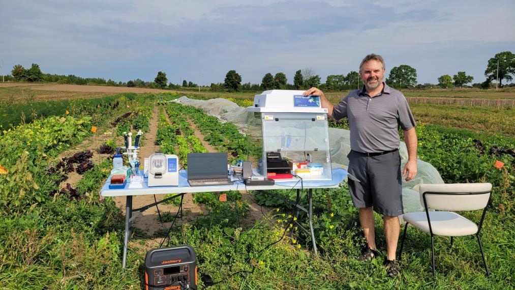 Chris Grainger stands in a green field wearing shorts and sandals, beside a table with scientific equipment on it. A large plexiglass box on the table contains smaller boxes and vials.
