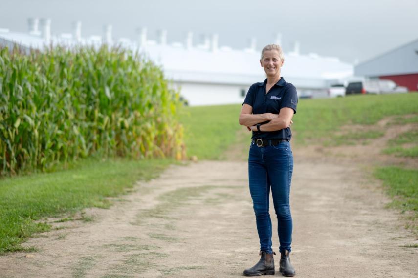 Terri poses, arms crossed and smiling, with the big barn of the swine research centre in the background