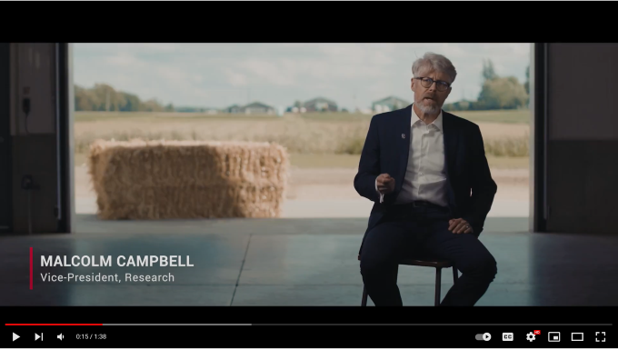 Dr. Malcolm Campbell, vice-president research, sits on a stool in a barn talking passionately. Hay is visible in the field outside the door to his back.