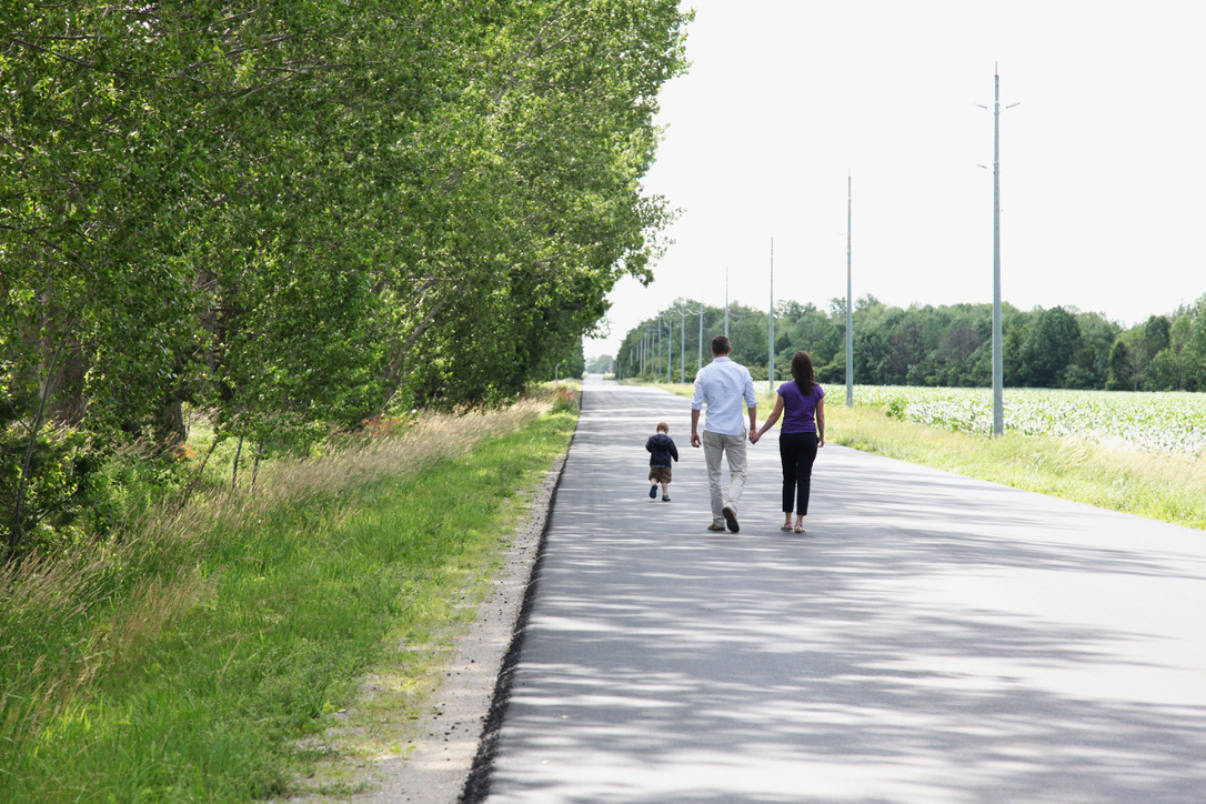A couple holding hands and their toddler walk down a road next to a field.