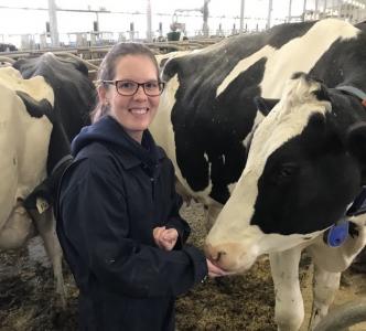 Patty Kedzierski, a young woman with glasses and a long ponytail, smiles beside several white and black dairy cows