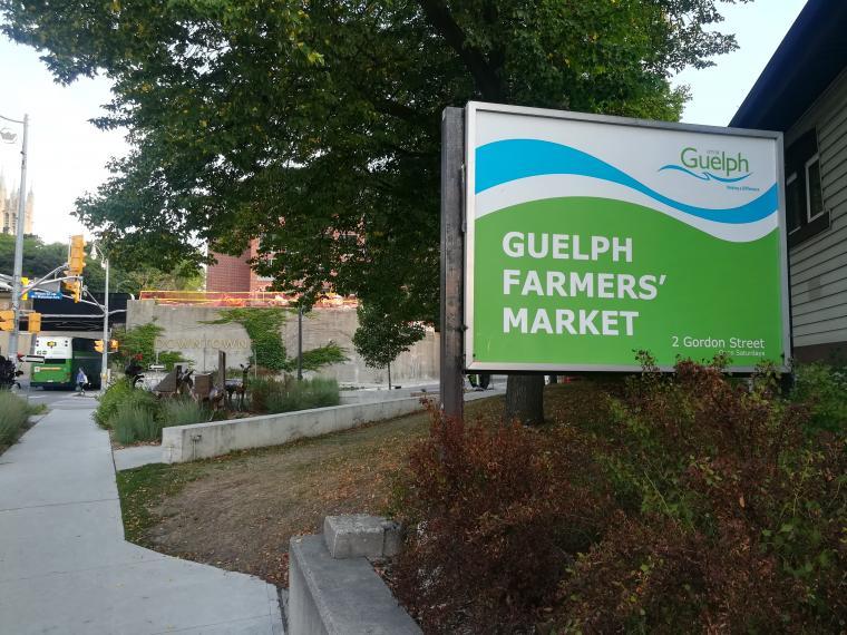 A photo of a sidewalk and path on the left, and a sign on the right that is blue, white and green. The text on the sign says, "Guelph Farmers Market". 