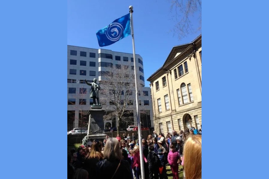 A crowd of people standing outside of buildings near a statue and a flag.