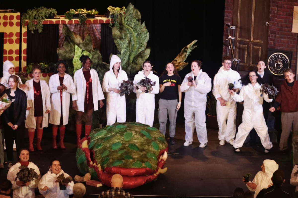 Dr. Kimberly Francis, professor and director, School of Fine Art and Music, College of Arts, University of Guelph, stands on stage at War Memorial Hall, holding a bouquet. Beside her is the "Little Shop of Horrors" student ensemble standing around Audrey II, the symbolic carnivorous plant from the production.