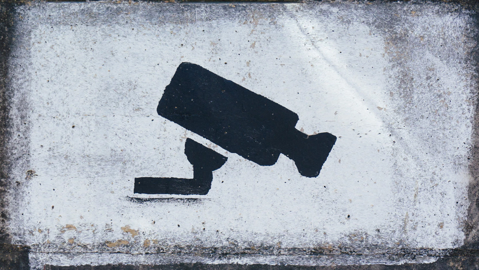 a surveillance camera, graffiti in black spray paint against a gray wall