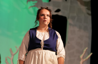 Karleigh Martin as The Baker’s Wife holding a lead for an animal and members of the ensemble on stage holding decorative tree branches in a production of "Into the Woods" at War Memorial Hall at the University of Guelph. The stage is set as a forest with trees. Photo credit: Joshua Lonuzzo 