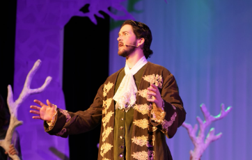 James Black as Rapunzel’s Prince and members of the ensemble on stage holding decorative tree branches in a production of "Into the Woods" at War Memorial Hall at the University of Guelph. The stage is set as a forest with trees. Photo credit: Joshua Lonuzzo  