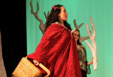  Charly Graham as Little Red Riding Hood and a member of the ensemble on stage holding decorative tree branches in a production of "Into the Woods" at War Memorial Hall at the University of Guelph. The stage is set as a forest with trees. Photo credit: Joshua Lonuzzo 